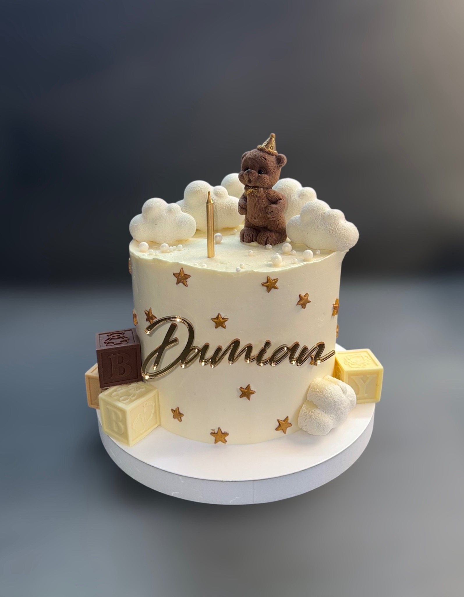 Tort urodzinowy z misiem, chmurkami i imieniem 'Damian', dekorowany gwiazdkami i kostkami z literami, na białym podeście, na szarym tle.