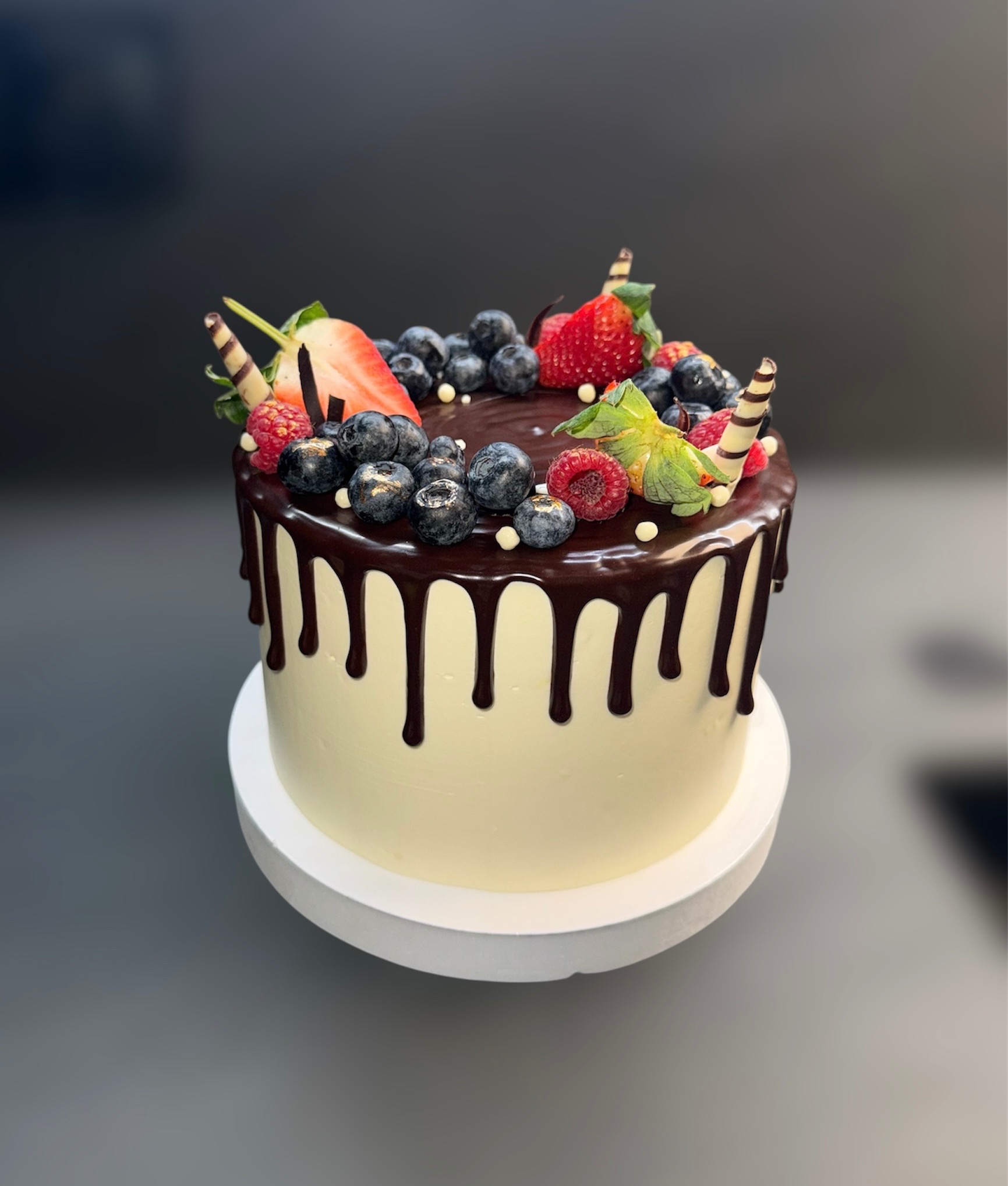 Tort z kremem, polewą czekoladową i świeżymi owocami: truskawkami, malinami i borówkami. Dekoracja z wafli. Całość na białym podeście, na ciemnym tle.