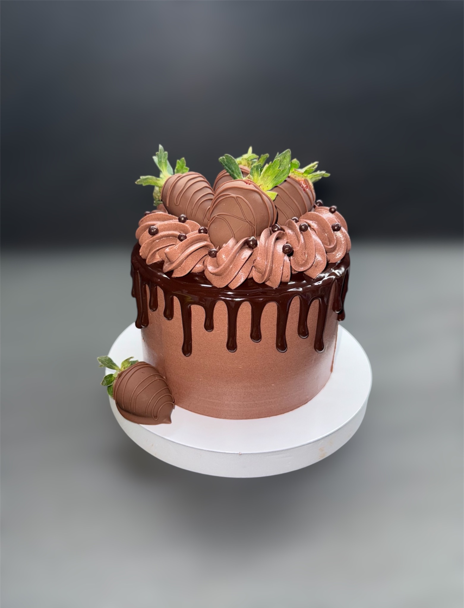 Czekoladowy tort z polewą i truskawkami w czekoladzie na białym podeście, na szarym tle. Dekoracja z kremu i kuleczek.