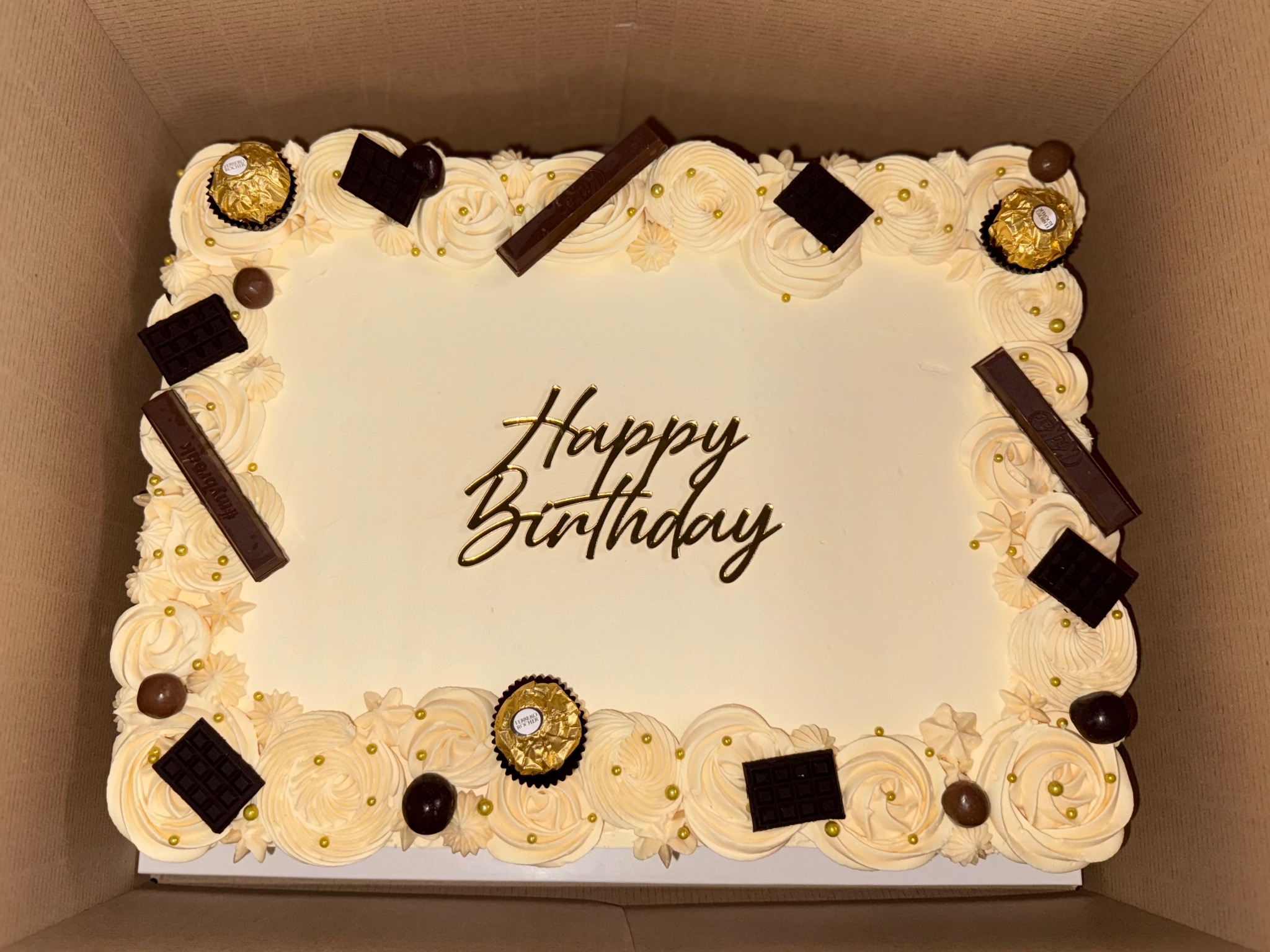 Prostokątny tort urodzinowy w kartonie, zdobiony kremem, czekoladkami, złotymi kuleczkami i napisem 'Happy Birthday' w kolorze złotym.