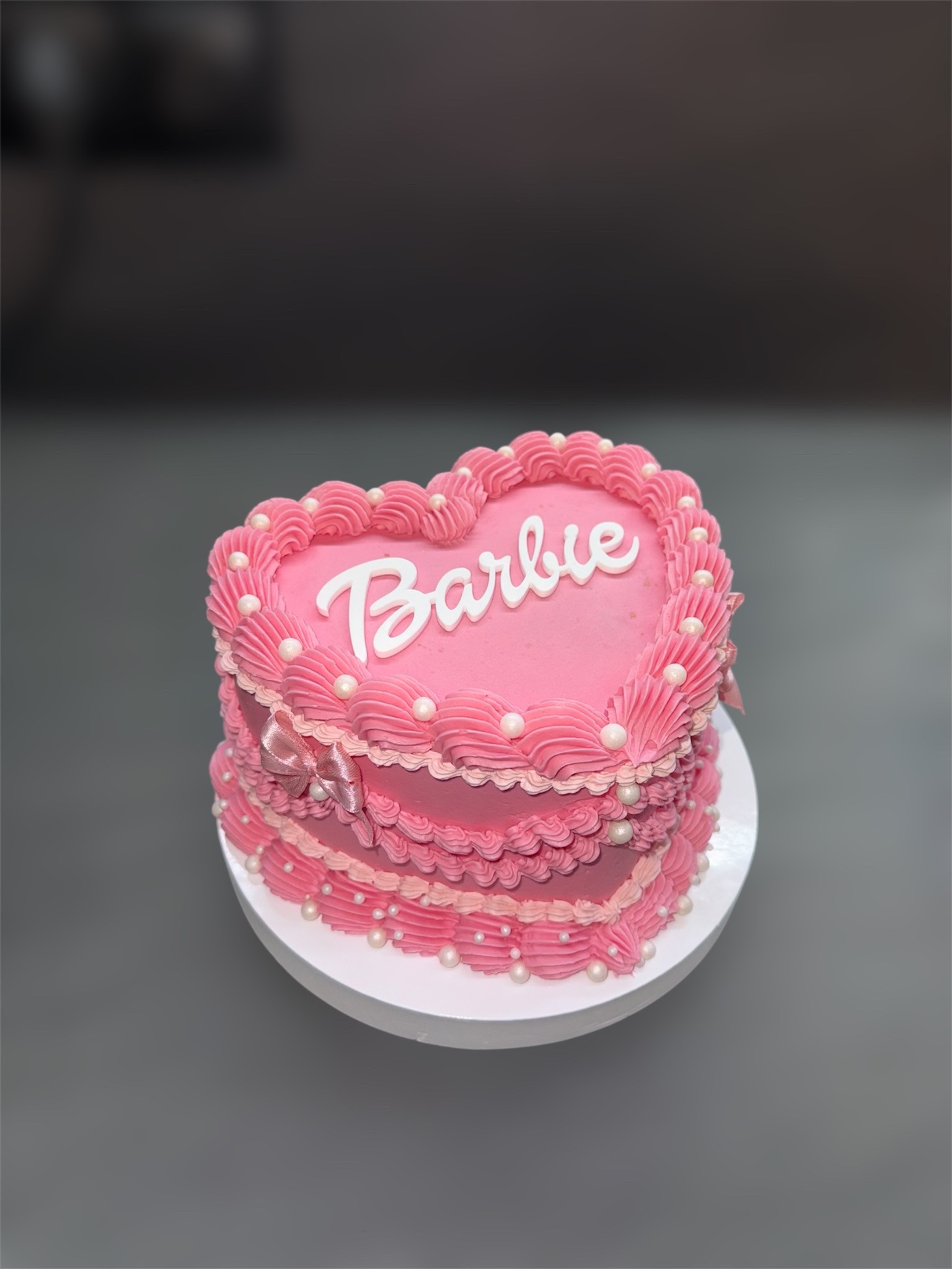 Różowy tort w kształcie serca z napisem 'Barbie', zdobiony kremem i perłami, na białym talerzu. Uroczy wypiek na specjalną okazję, z delikatną wstążką.