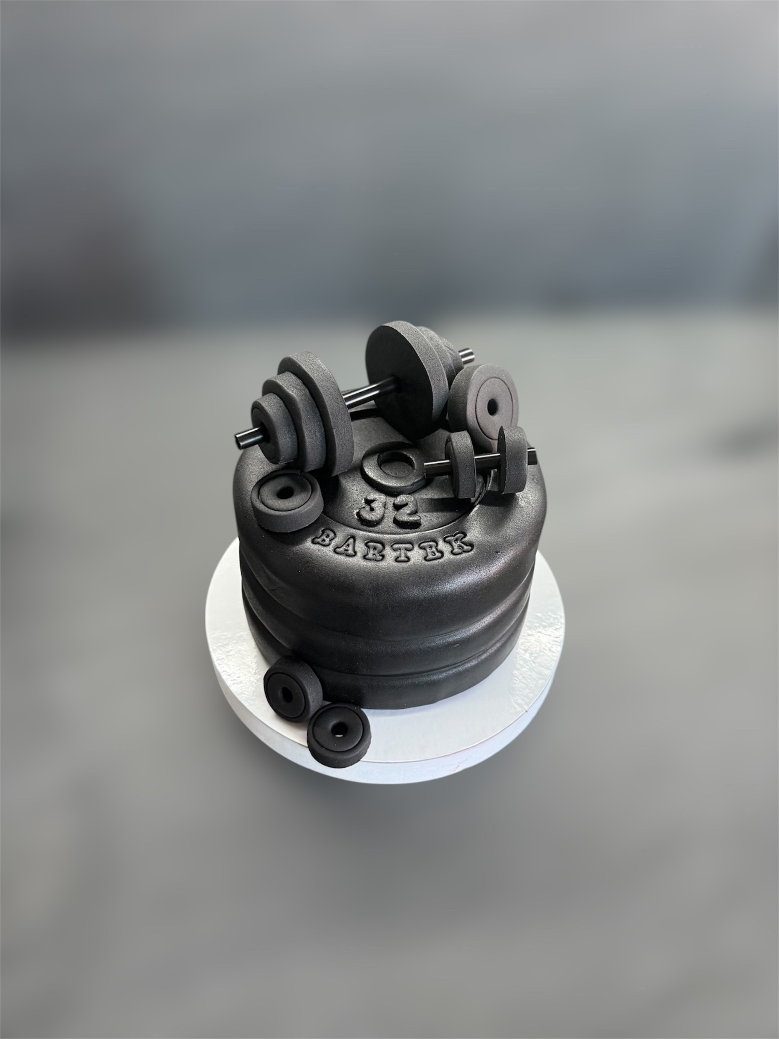Czarny tort z motywem siłowni: sztangi i ciężary. Napis '32 Bartek'. Idealny dla fana fitnessu. Minimalistyczne tło, nowoczesny design.