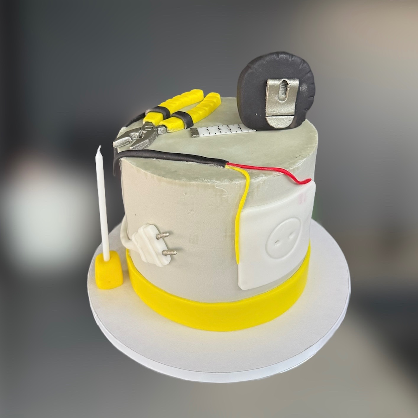 Tematyczny tort w kształcie walca, dekorowany elementami instalacji elektrycznej: gniazdko, wtyczka, przewody, miernik i kombinerki. Żółte akcenty.