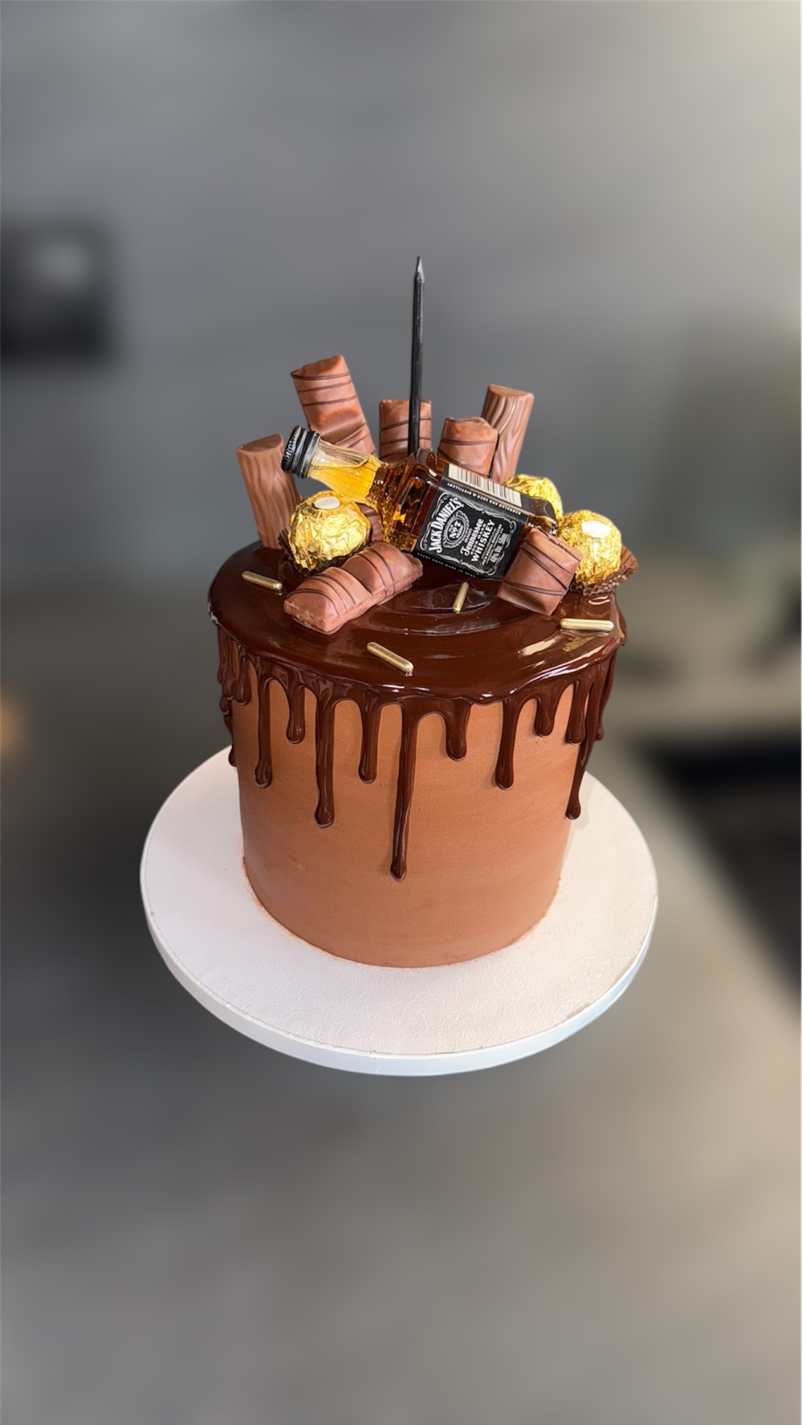 Czekoladowy tort z polewą, dekorowany batonikami, złotymi pralinkami i miniaturową butelką alkoholu. Na górze pojedyncza świeczka.