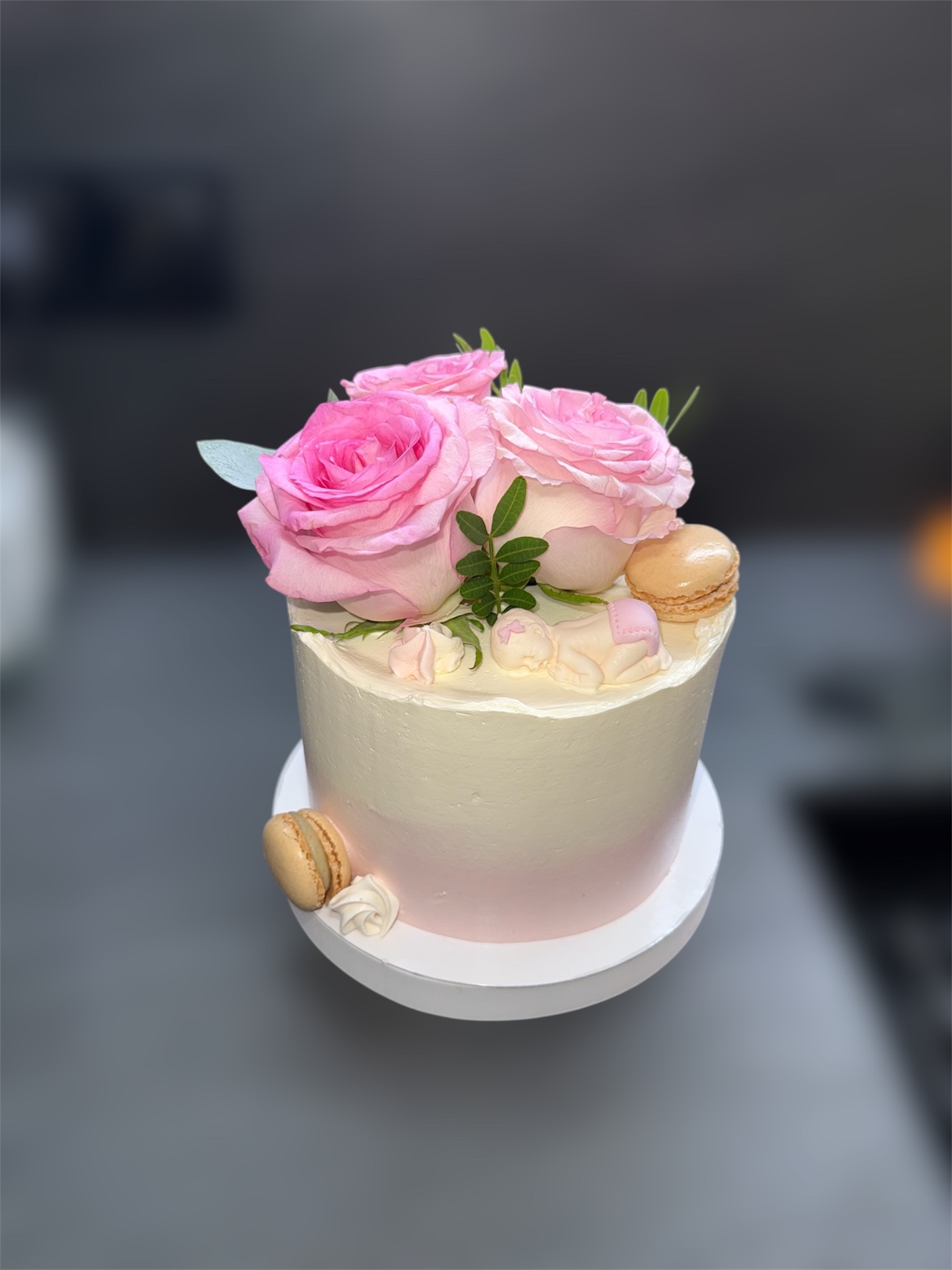 Tort zdobiony różowymi różami, makaronikami i figurką dziecka, na białym podeście. Kremowy tort z delikatnym różowym ombre u dołu. Ujęcie z góry, na szarym tle.