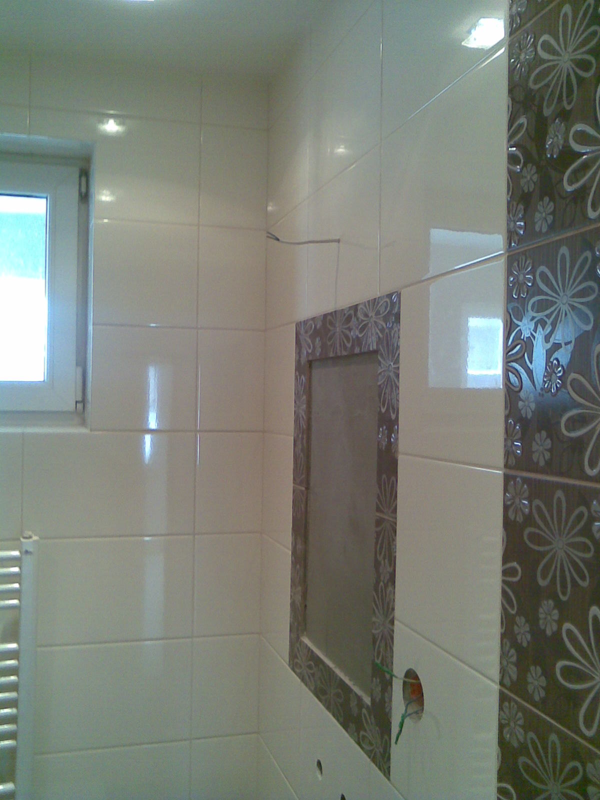 Białe, błyszczące płytki ścienne w łazience z dekoracyjnym pasem z motywem kwiatowym i otworem rewizyjnym z ramą również w kwiaty, widoczne okno i grzejnik.