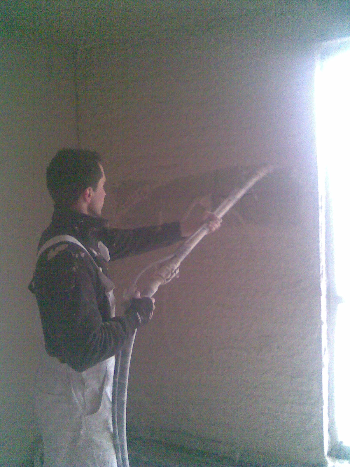 Pracownik w kombinezonie natryskuje maszynowo zaprawę na ścianę wewnątrz budynku, widoczne przewody doprowadzające materiał.