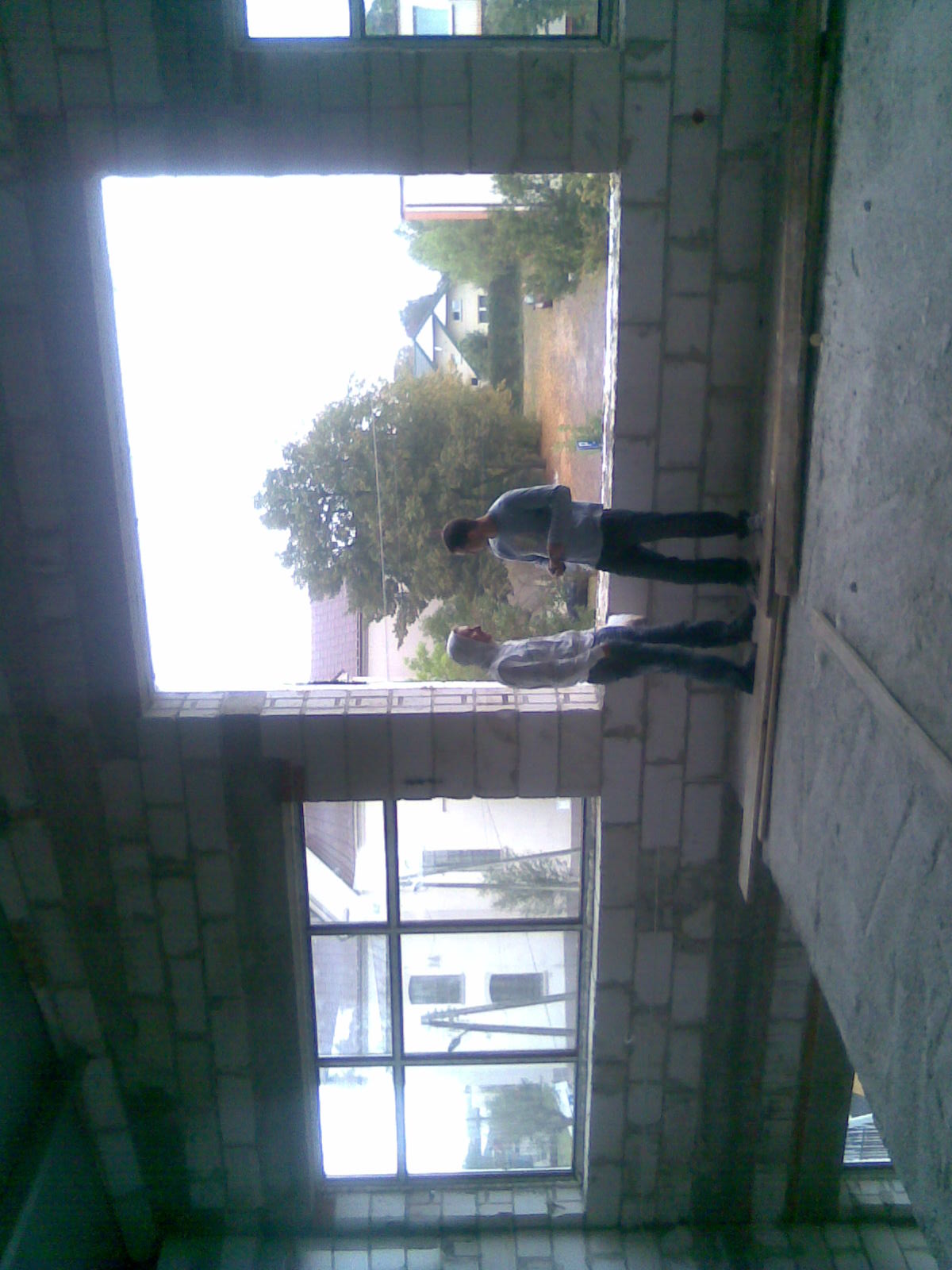 Dwóch mężczyzn w surowym wnętrzu budynku w trakcie tynkowania maszynowego ściany z pustaków, widok przez okno na zewnątrz budynku.