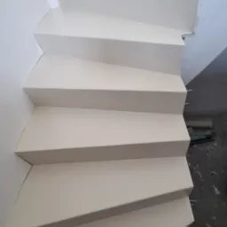 Białe schody wewnętrzne z wykończeniem, widok z dołu, zarys instalacji elektrycznej na ścianie, częściowo widoczne narzędzia na podłodze.