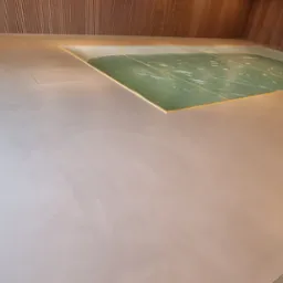 Jasna, gładka posadzka w pomieszczeniu z drewnianym wykończeniem ścian. Widoczny fragment basenu z zieloną wodą i złotym obramowaniem. Minimalistyczny design.