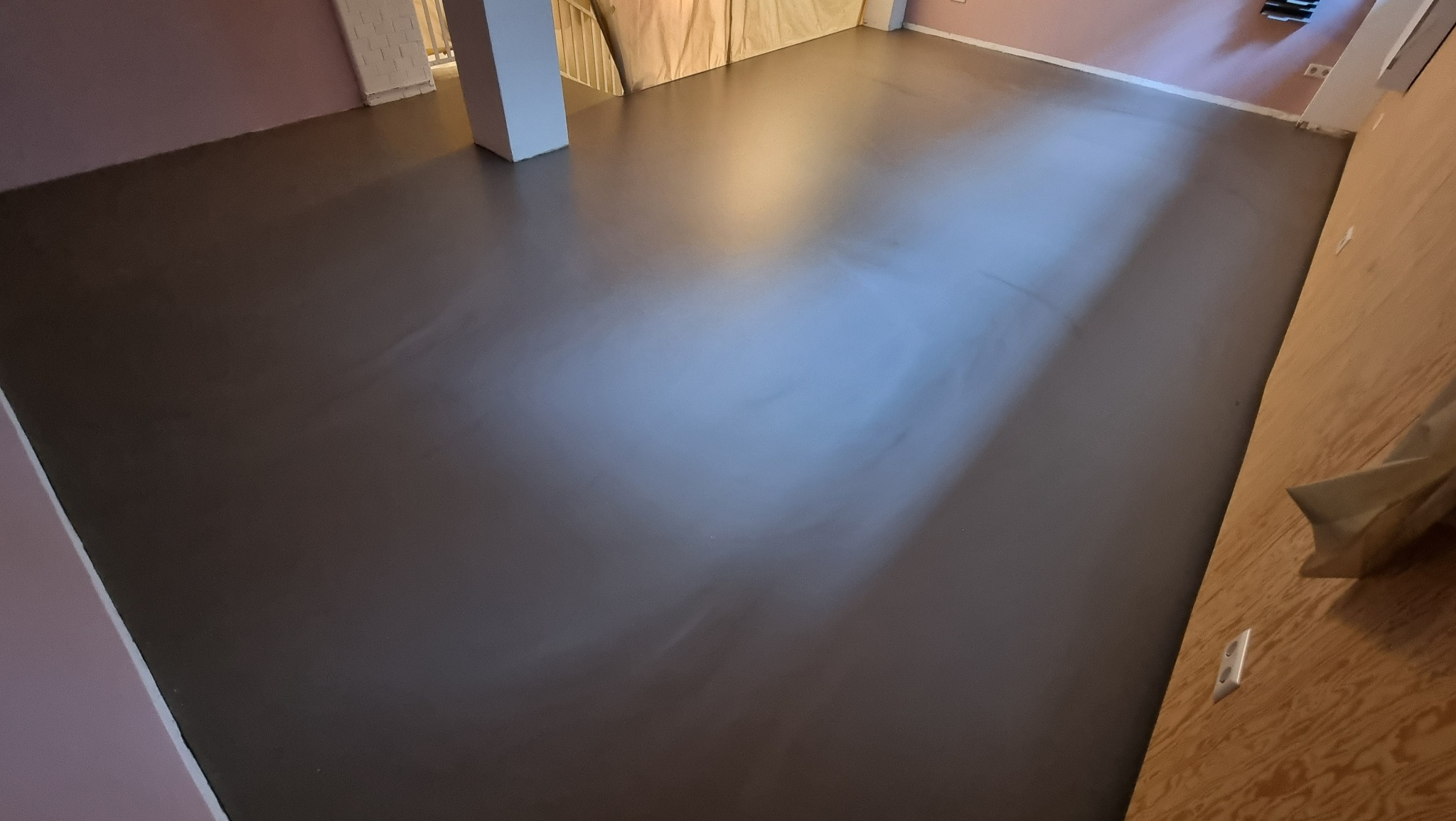 Gładka, szara posadzka w jasnym pomieszczeniu z kolumną i schodami w tle. Nowoczesne wykończenie podłogi, widoczne detale ścian i oświetlenie.