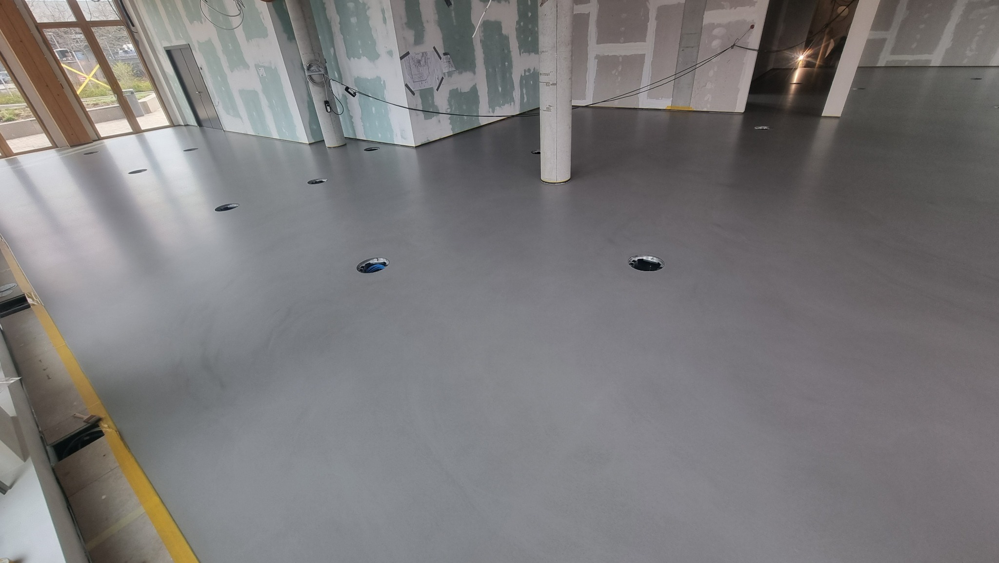 Szara, gładka podłoga w surowym wnętrzu z widocznymi otworami na instalacje i żółtą taśmą zabezpieczającą przy ścianie. W tle ściany przygotowane do wykończenia.