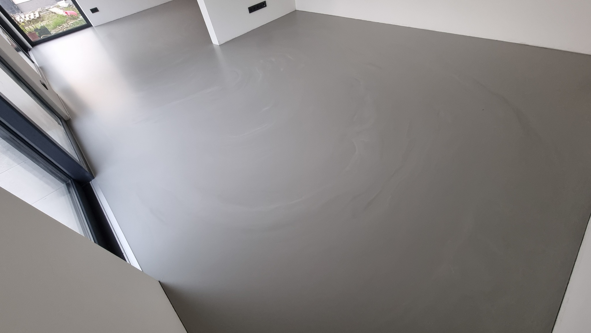 Szara, gładka posadzka w jasnym wnętrzu z dużym oknem balkonowym. Widoczny subtelny wzór na powierzchni. Minimalistyczny design.