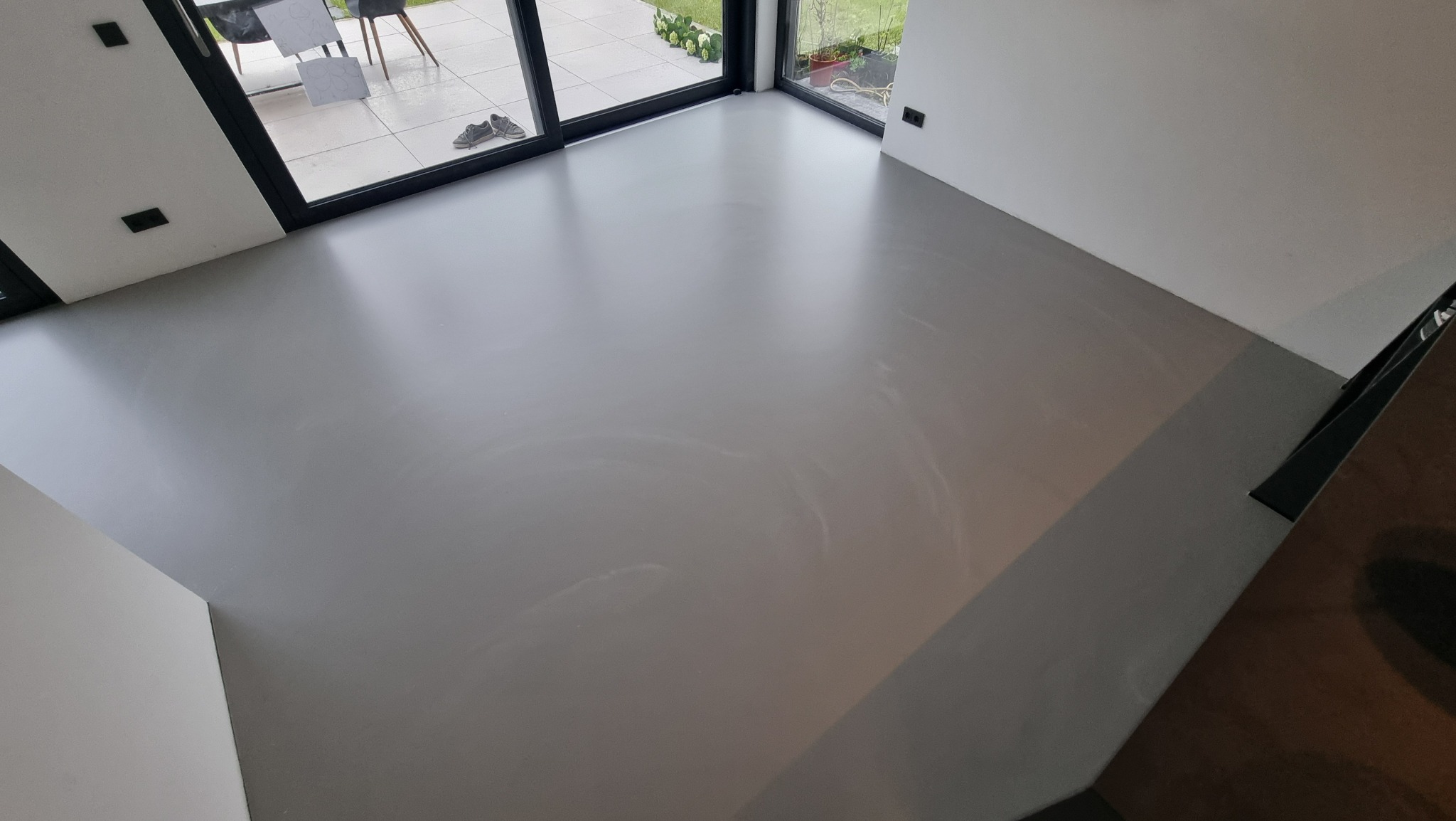 Gładka, szara podłoga w nowoczesnym wnętrzu z dużymi oknami. Widoczne subtelne wzory na powierzchni posadzki i para butów przy oknie.
