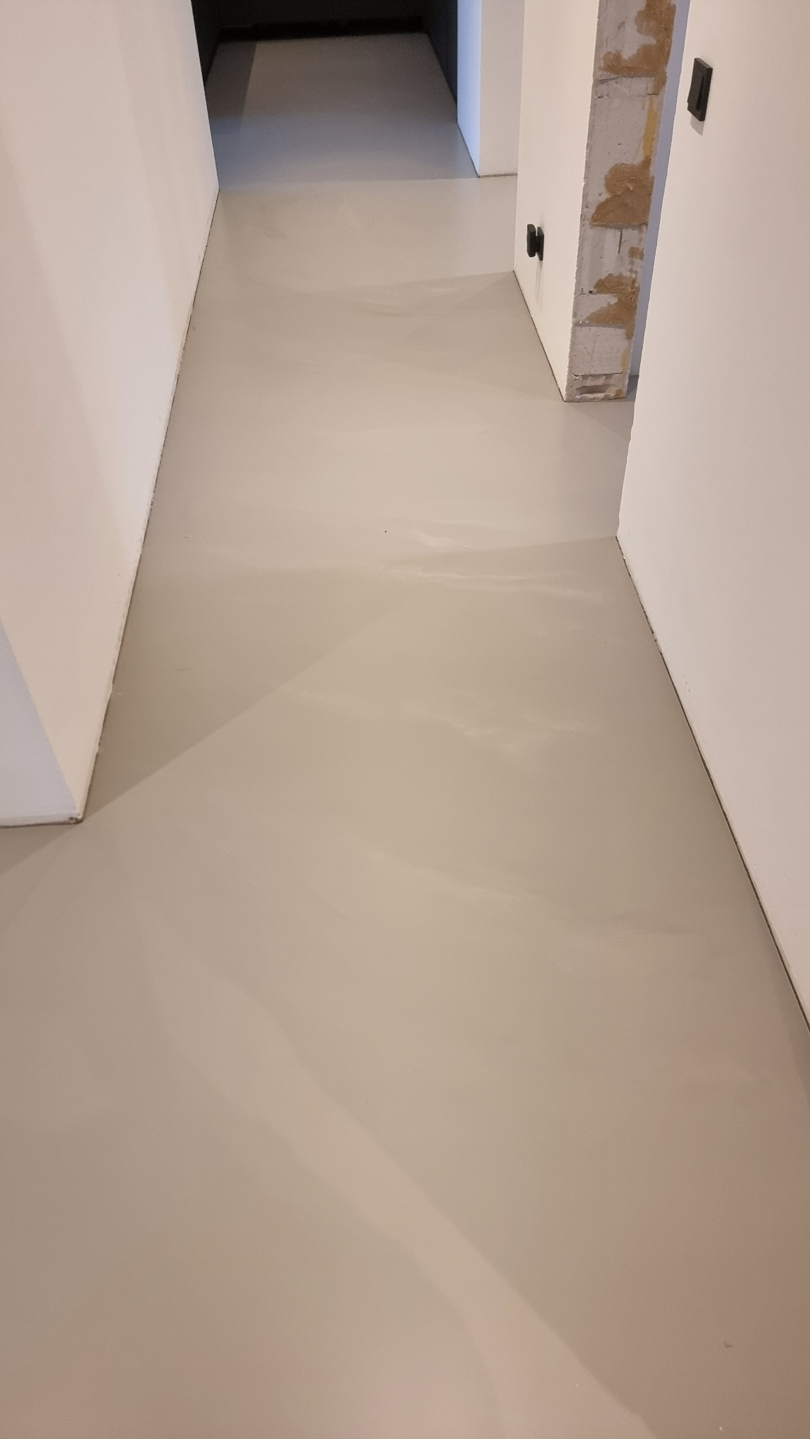 Jasnoszara, gładka posadzka żywiczna w korytarzu z białymi ścianami. Widoczny fragment surowego muru przy drzwiach.