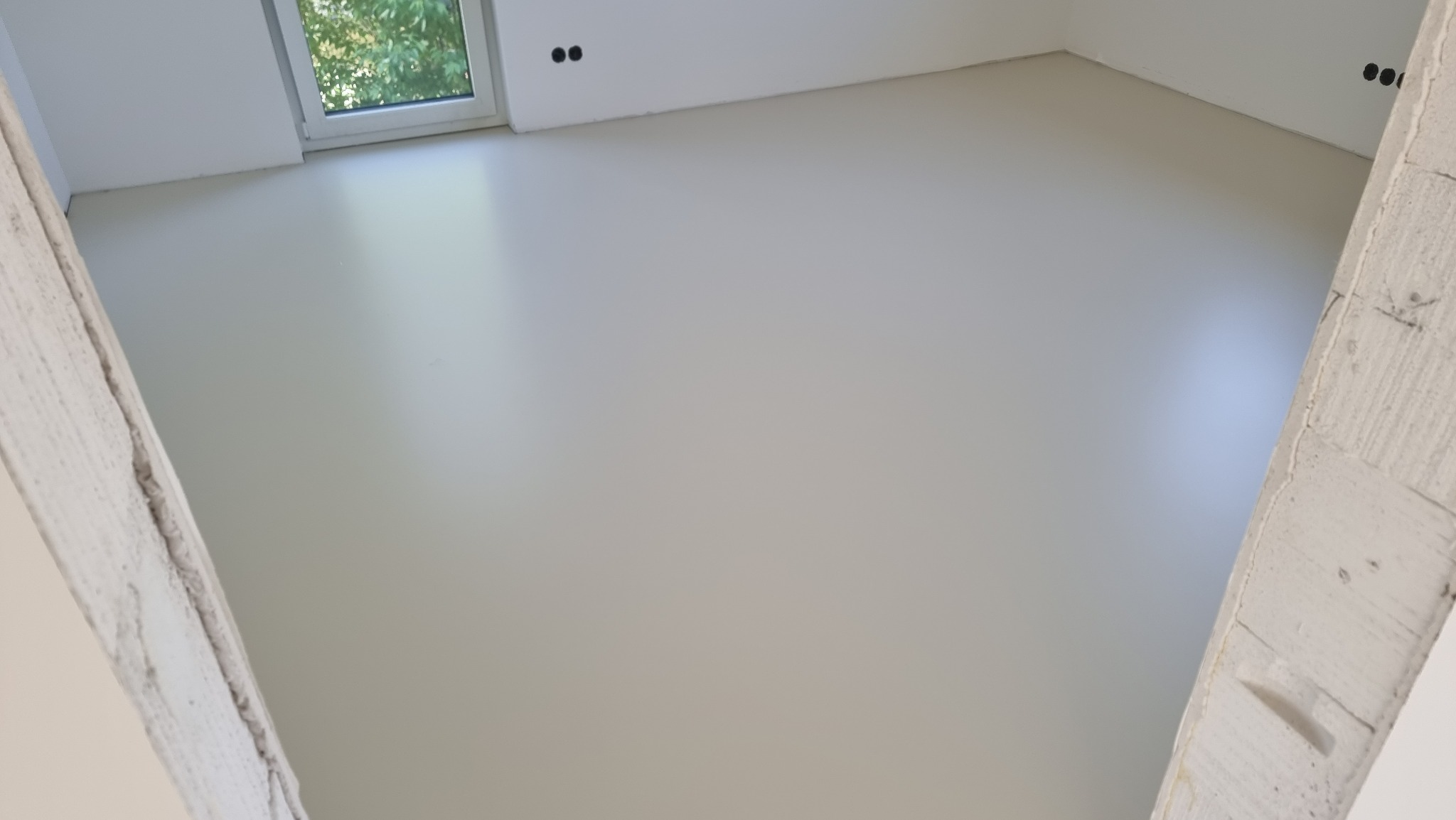 Jasna, jednolita posadzka w nowym wnętrzu. Świeżo wylana, gładka powierzchnia w surowym stanie, widoczne ślady po szalunku na ścianach. Minimalistyczny design.