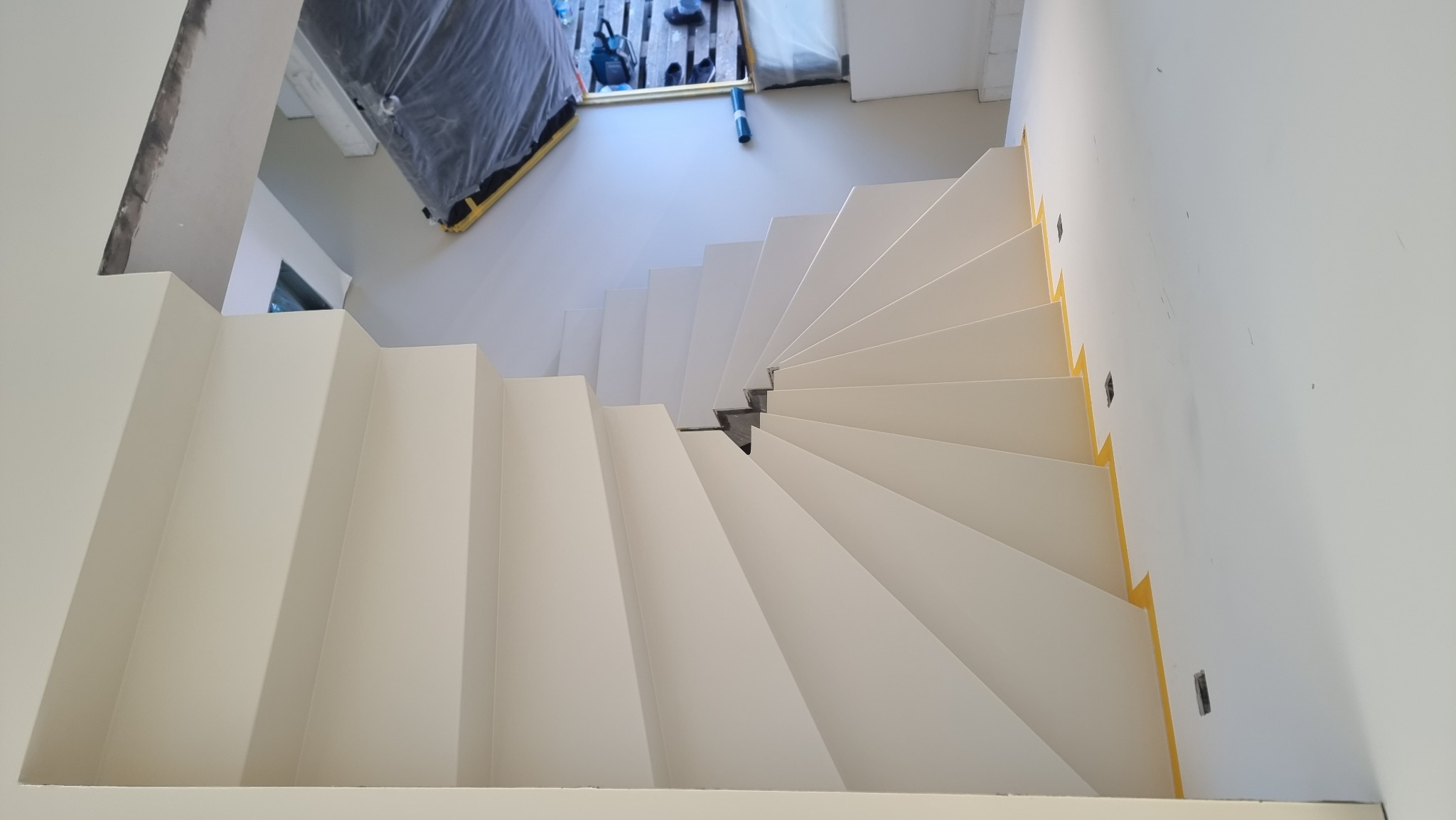 Białe schody spiralne z widocznymi zabezpieczeniami taśmą malarską, ujęcie z góry. Widoczne niedoskonałości wykończenia ścian.