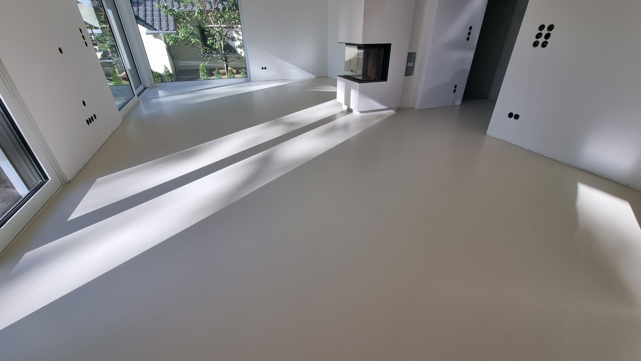 Jasna, gładka posadzka w minimalistycznym wnętrzu z kominkiem. Światło słoneczne tworzy geometryczne wzory na podłodze. Widoczne okno z zielenią na zewnątrz.