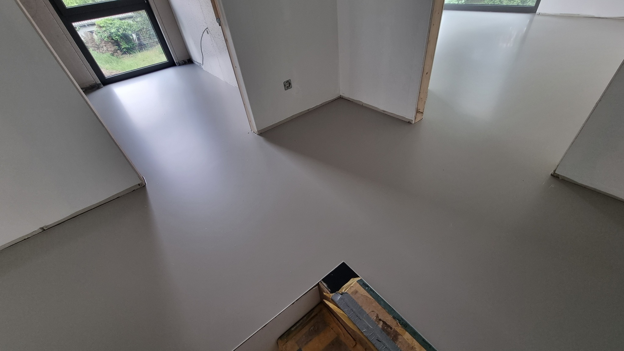 Jasnoszara, gładka posadzka w nowym budynku z widocznym fragmentem niezabezpieczonej podłogi. Minimalistyczne wnętrze z oknem i surowymi ścianami.