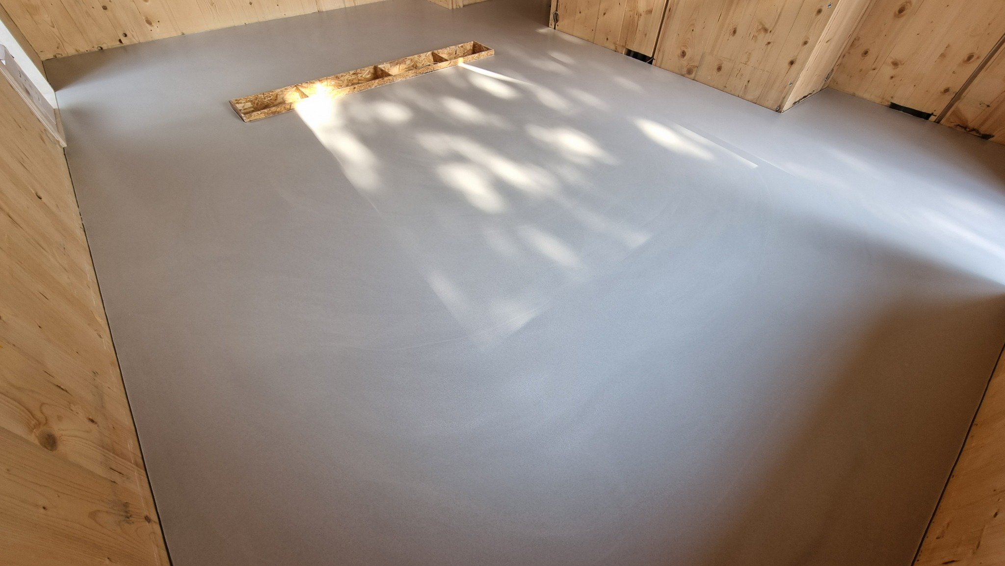 Gładka, szara powierzchnia w pomieszczeniu z drewnianymi ścianami. Na podłodze widoczne refleksy światła i drewniana listwa przypodłogowa.