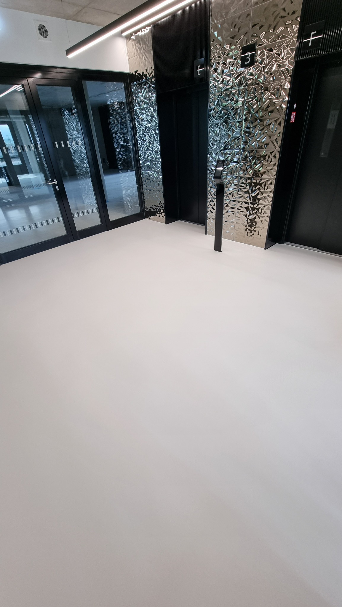 Jasna, jednolita posadzka w nowoczesnym budynku biurowym, z widocznymi drzwiami i windami o metalicznych, teksturowanych ścianach.