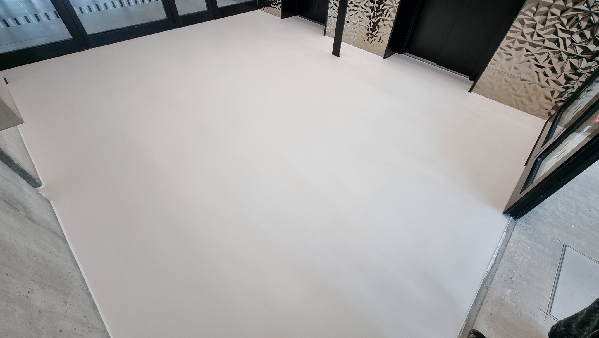 Jasna, jednolita posadzka w komercyjnym wnętrzu, widok z góry. Minimalistyczny design, szklane drzwi wejściowe, metalowe panele dekoracyjne na ścianach.