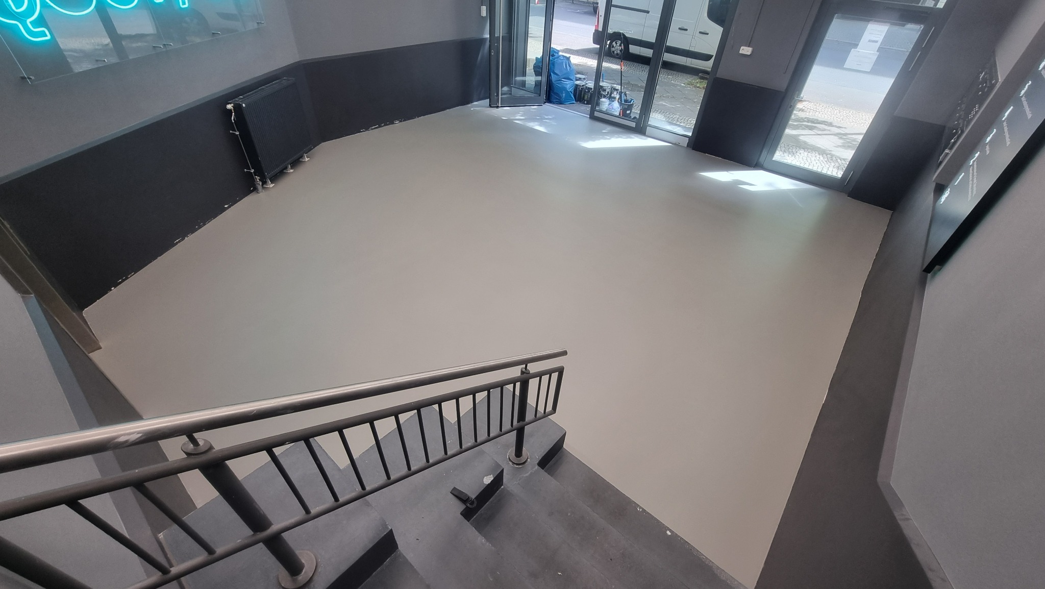 Jasnoszara, gładka posadzka w lokalu z widocznymi schodami i metalową barierką. Przez otwarte drzwi widać zaparkowany samochód dostawczy.