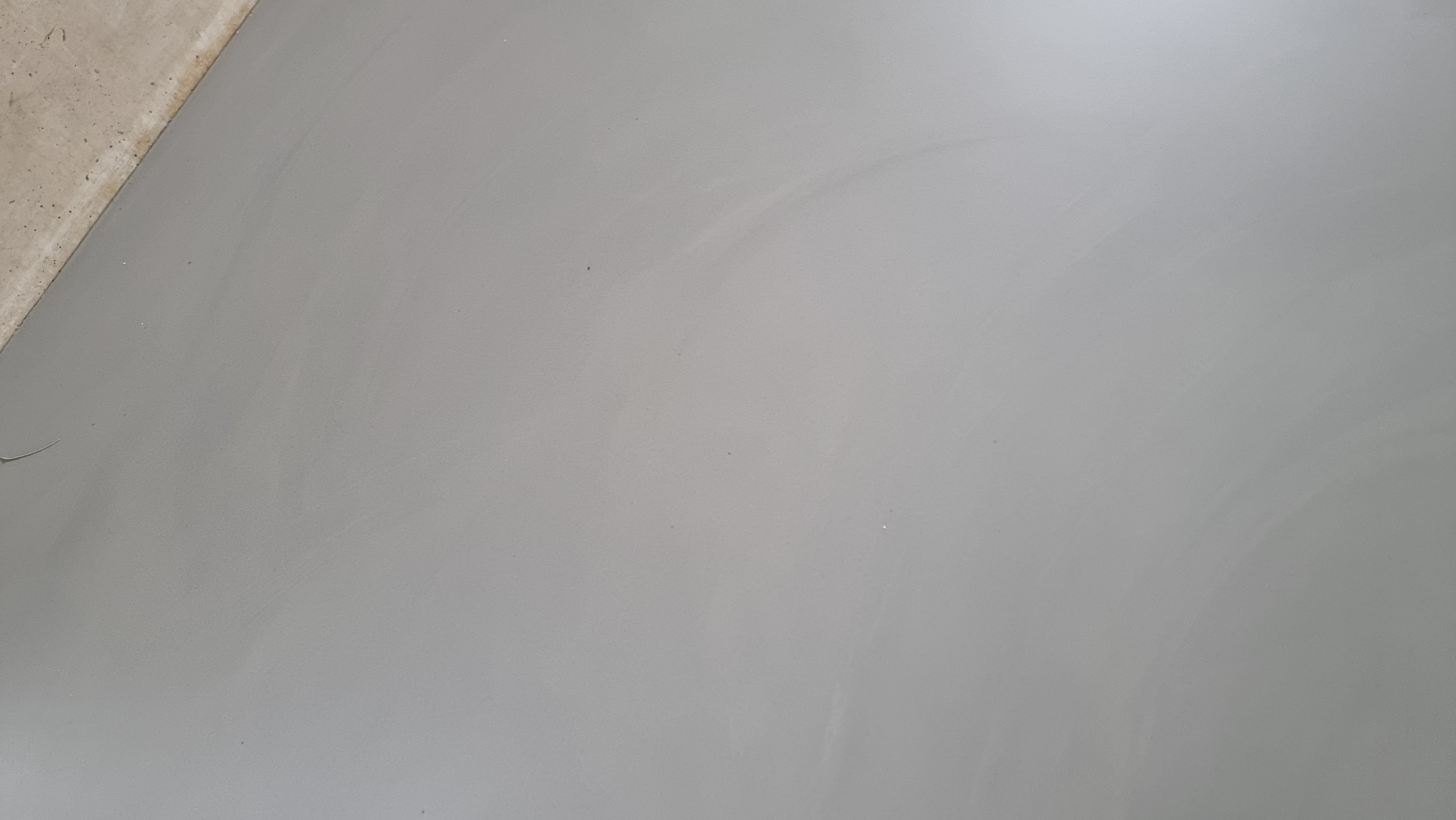 Gładka, szara powierzchnia posadzki z delikatnym połyskiem, widoczny fragment ściany w lewym górnym rogu. Minimalistyczne ujęcie, jednolite tło.