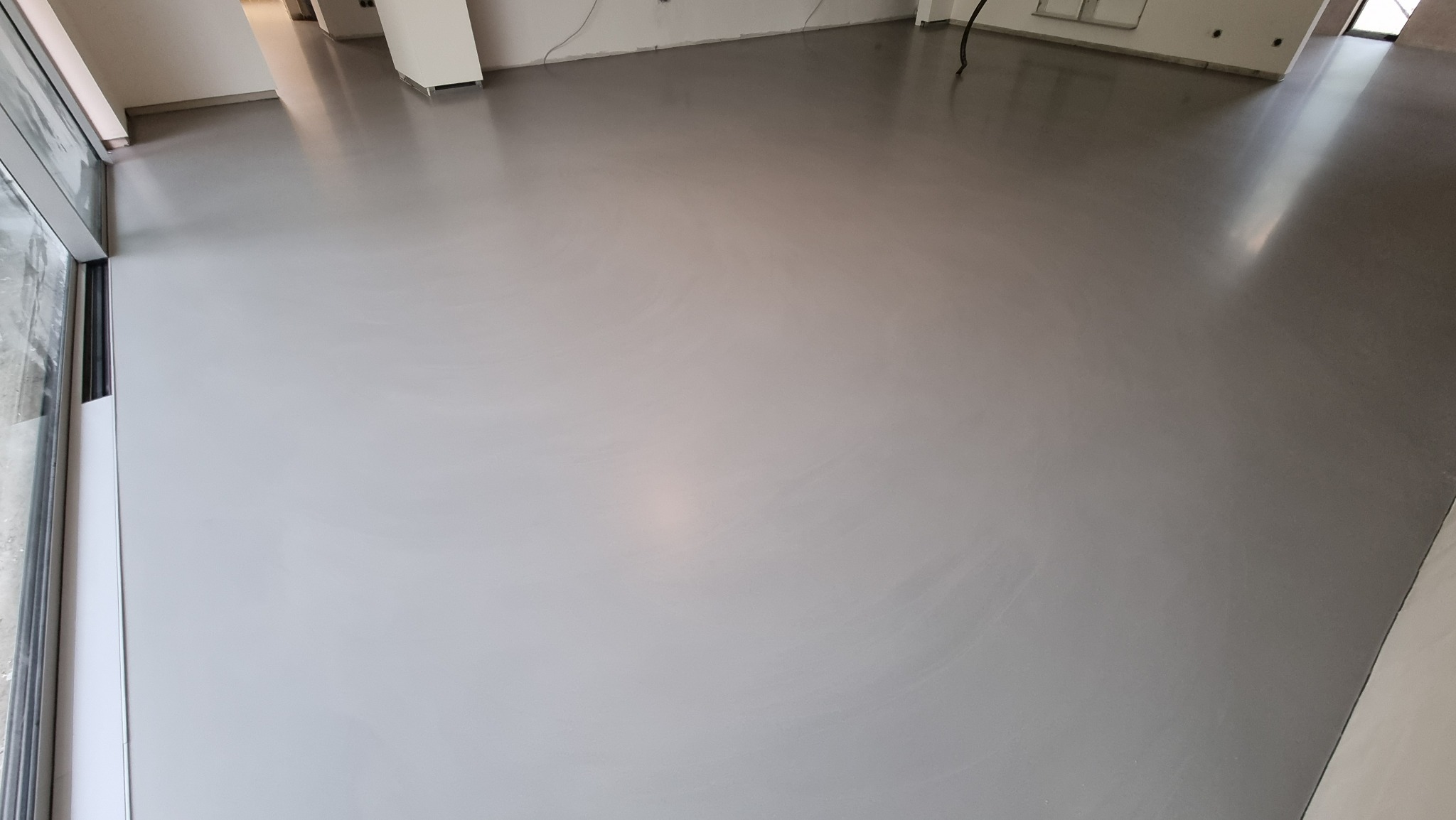 Jasnoszara, gładka posadzka żywiczna w nowym, pustym pomieszczeniu z dużymi oknami. Widoczny fragment ściany i instalacje elektryczne.