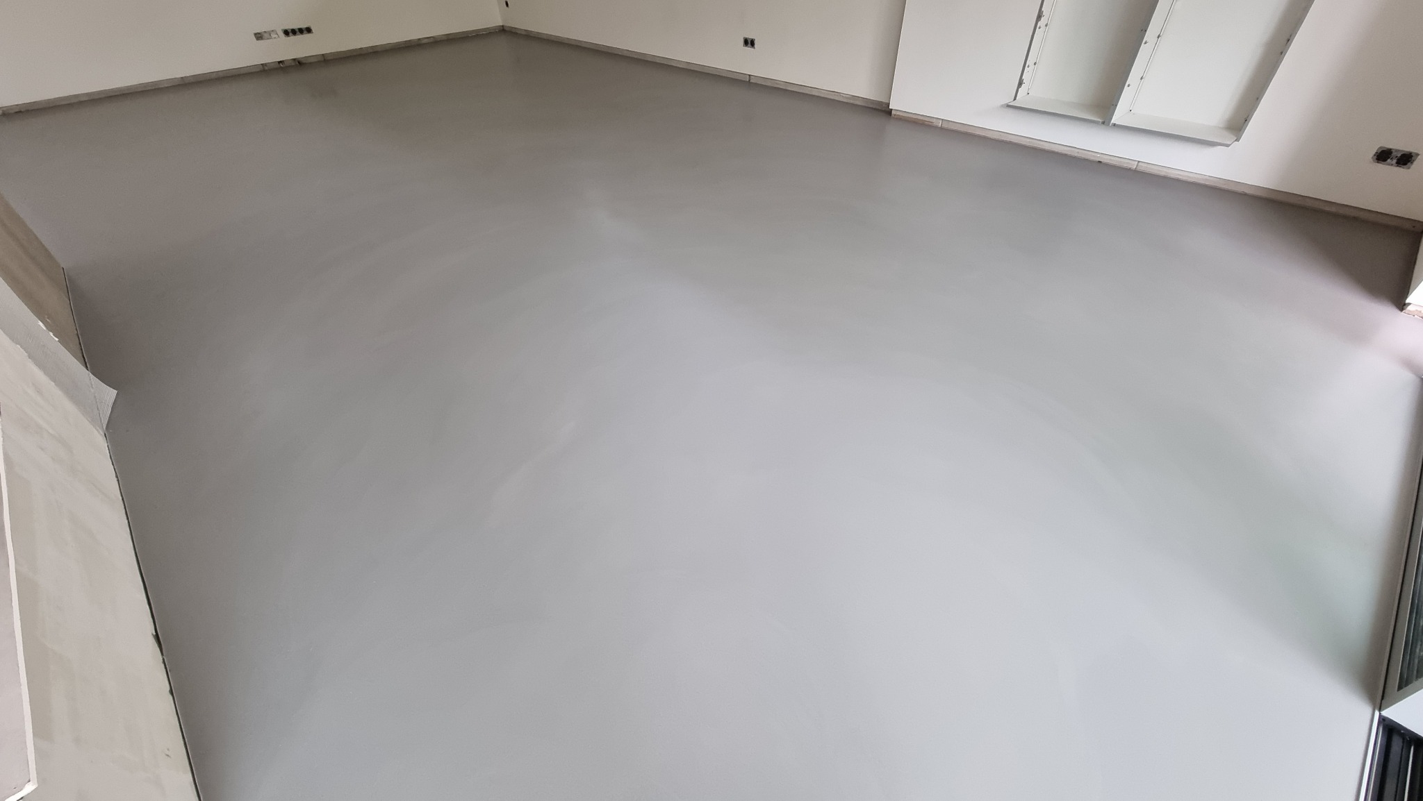 Jasnoszara, gładka posadzka w nowym wnętrzu, widok z góry. Minimalistyczny styl, świeżo wylana powierzchnia. Widoczne gniazdka elektryczne na ścianie.