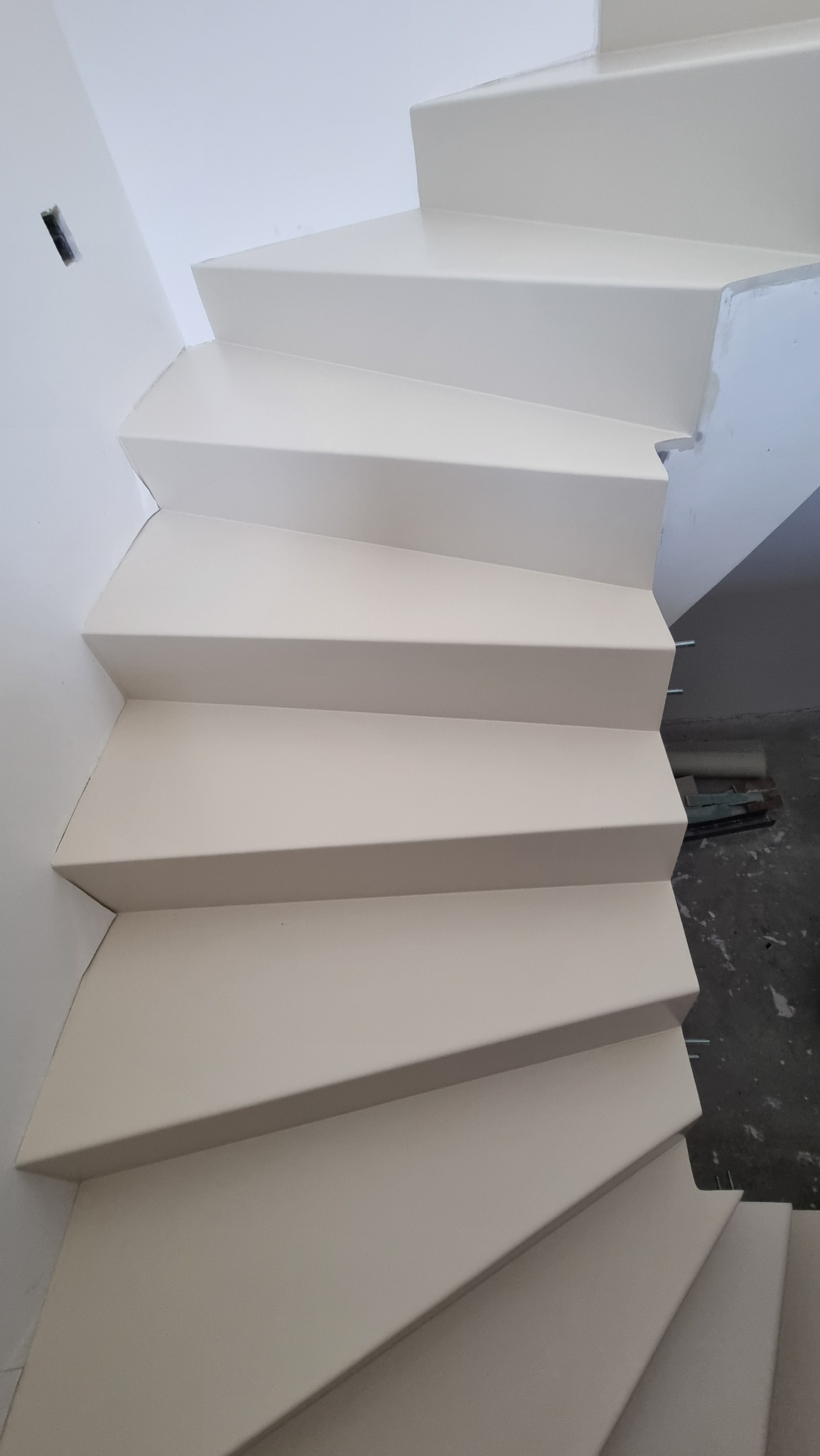 Białe schody wewnętrzne z wykończeniem, widok z dołu, zarys instalacji elektrycznej na ścianie, częściowo widoczne narzędzia na podłodze.