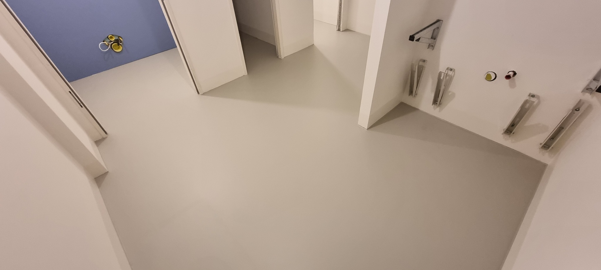 Jasnoszara, gładka podłoga w korytarzu z widocznymi instalacjami na ścianie. Ujęcie z góry, perspektywa wnętrza z otwartymi drzwiami do innych pomieszczeń.