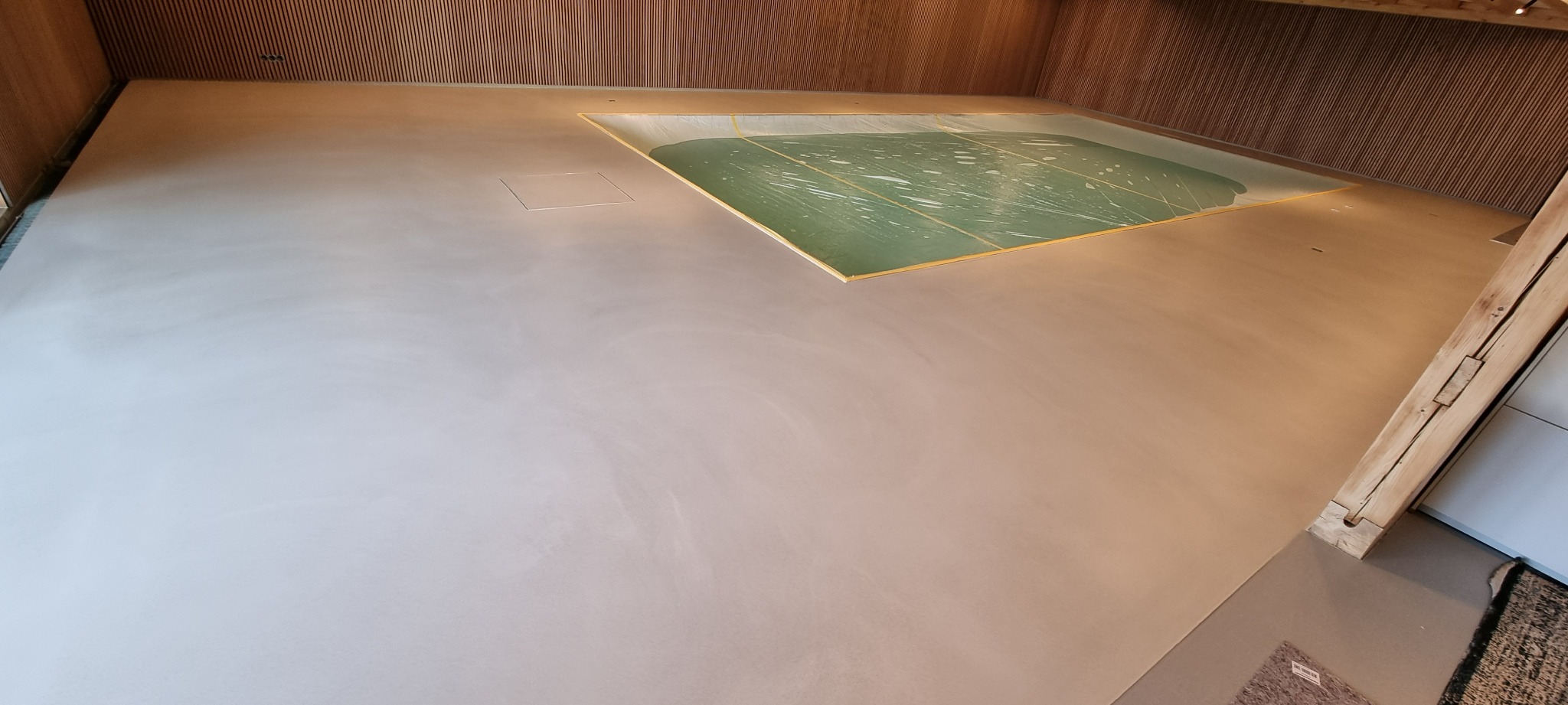 Jasna, gładka posadzka w pomieszczeniu z drewnianym wykończeniem ścian. Widoczny fragment basenu z zieloną wodą i złotym obramowaniem. Minimalistyczny design.