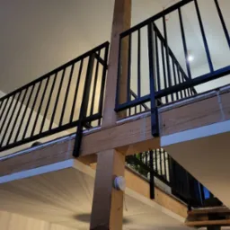 Nowoczesna, czarna balustrada na antresoli z drewnianymi elementami konstrukcyjnymi, widziana z dołu, z widoczną lampą wiszącą i fragmentem schodów.