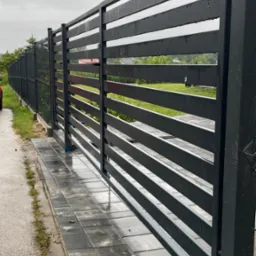 Nowoczesna, pozioma, metalowa brama przesuwna w kolorze grafitowym, zamontowana na posesji z kostką brukową. Widok z bliska, w tle zieleń ogrodu.