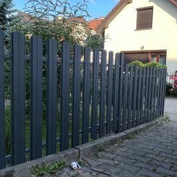 Nikodem Liszka - Grafitowe, metalowe ogrodzenie sztachetowe przed domem, na kostce brukowej leżą przewody. Tło: dom z brązową roletą i zielona roślinność.
