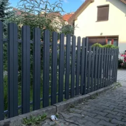 Grafitowe, metalowe ogrodzenie sztachetowe przed domem, na kostce brukowej leżą przewody. Tło: dom z brązową roletą i zielona roślinność.