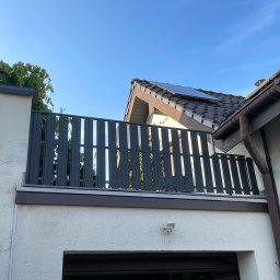 Nikodem Liszka - Nowoczesna balustrada balkonowa z pionowych paneli w Trzebini. Szara konstrukcja na białym budynku, widok z dołu na tle błękitnego nieba i fragmentu dachu.