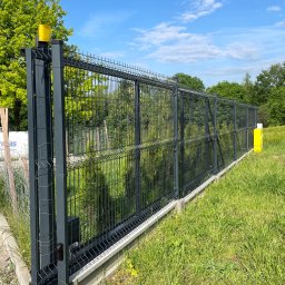 Nikodem Liszka - Przesuwna brama panelowa w kolorze grafitowym, z żółtą lampą ostrzegawczą, zamontowana na betonowej podstawie. Widoczne zielone trawniki i drzewa w tle.