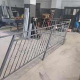 Nikodem Liszka - Szara, metalowa balustrada w warsztacie. Widoczne spawy i surowy metal. W tle narzędzia i materiały, co sugeruje etap produkcji lub montażu.