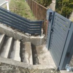 Nikodem Liszka - Nowoczesna, grafitowa furtka z poziomymi szczeblami, zamontowana przy betonowych schodach i ogrodzeniu z siatki. Ujęcie z dołu, widok na otwartą furtkę.