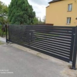 Nikodem Liszka - Nowoczesna, przesuwna brama wjazdowa z poziomych, grafitowych profili, na tle zieleni i żółtego domu. Widoczne detale konstrukcyjne i mechanizm bramy.