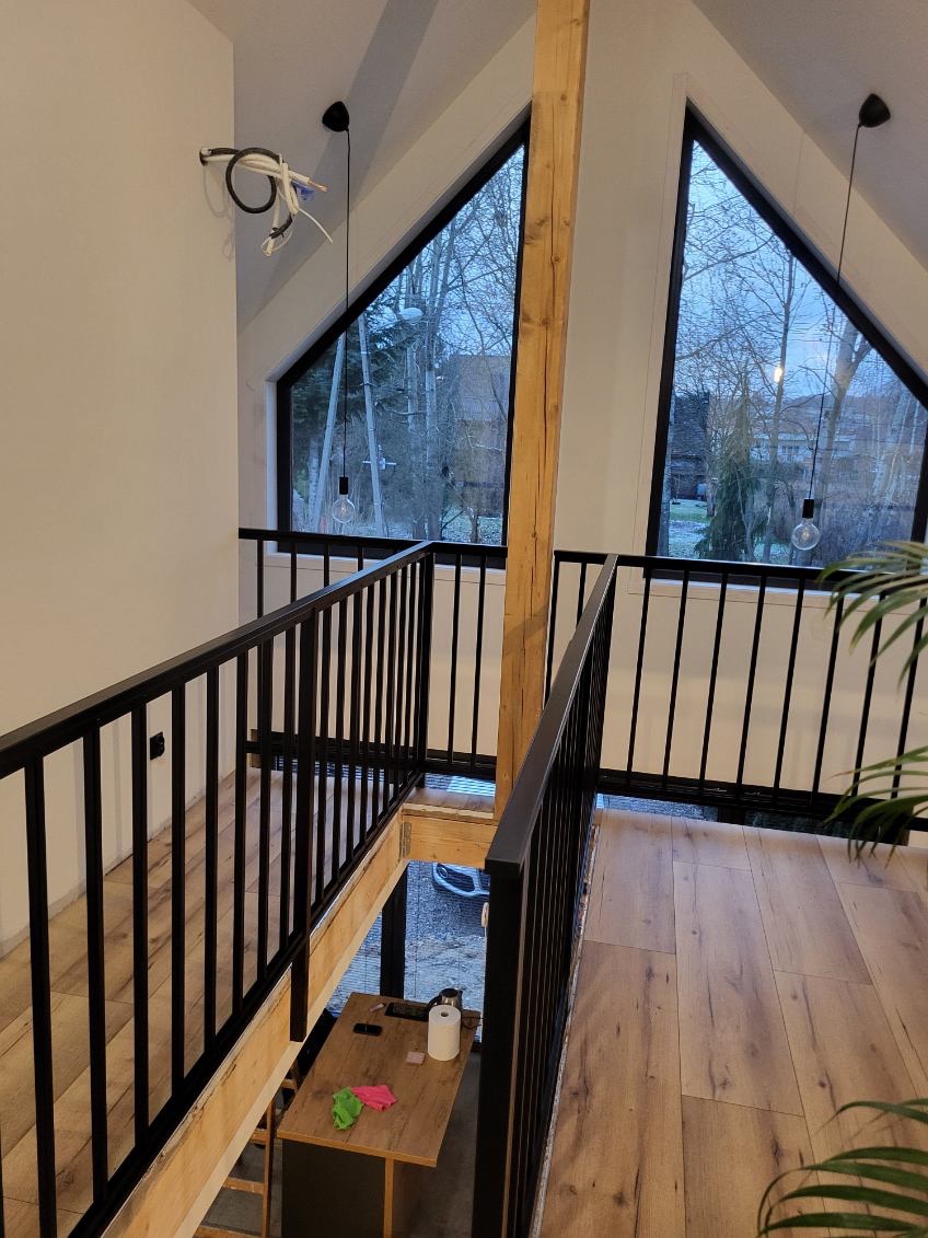 Nowoczesna balustrada schodowa z czarnych, metalowych prętów w jasnym wnętrzu z drewnianą podłogą i dużymi oknami trójkątnymi.