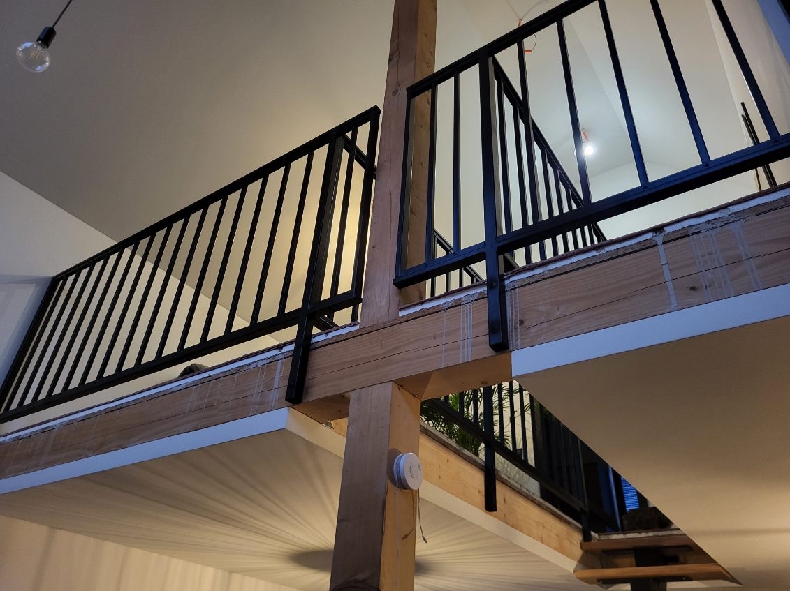 Nowoczesna, czarna balustrada na antresoli z drewnianymi elementami konstrukcyjnymi, widziana z dołu, z widoczną lampą wiszącą i fragmentem schodów.