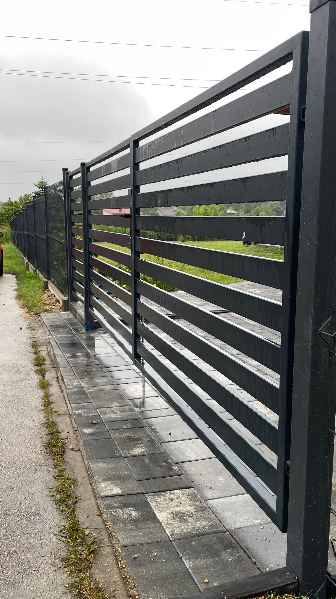 Nowoczesna, pozioma, metalowa brama przesuwna w kolorze grafitowym, zamontowana na posesji z kostką brukową. Widok z bliska, w tle zieleń ogrodu.