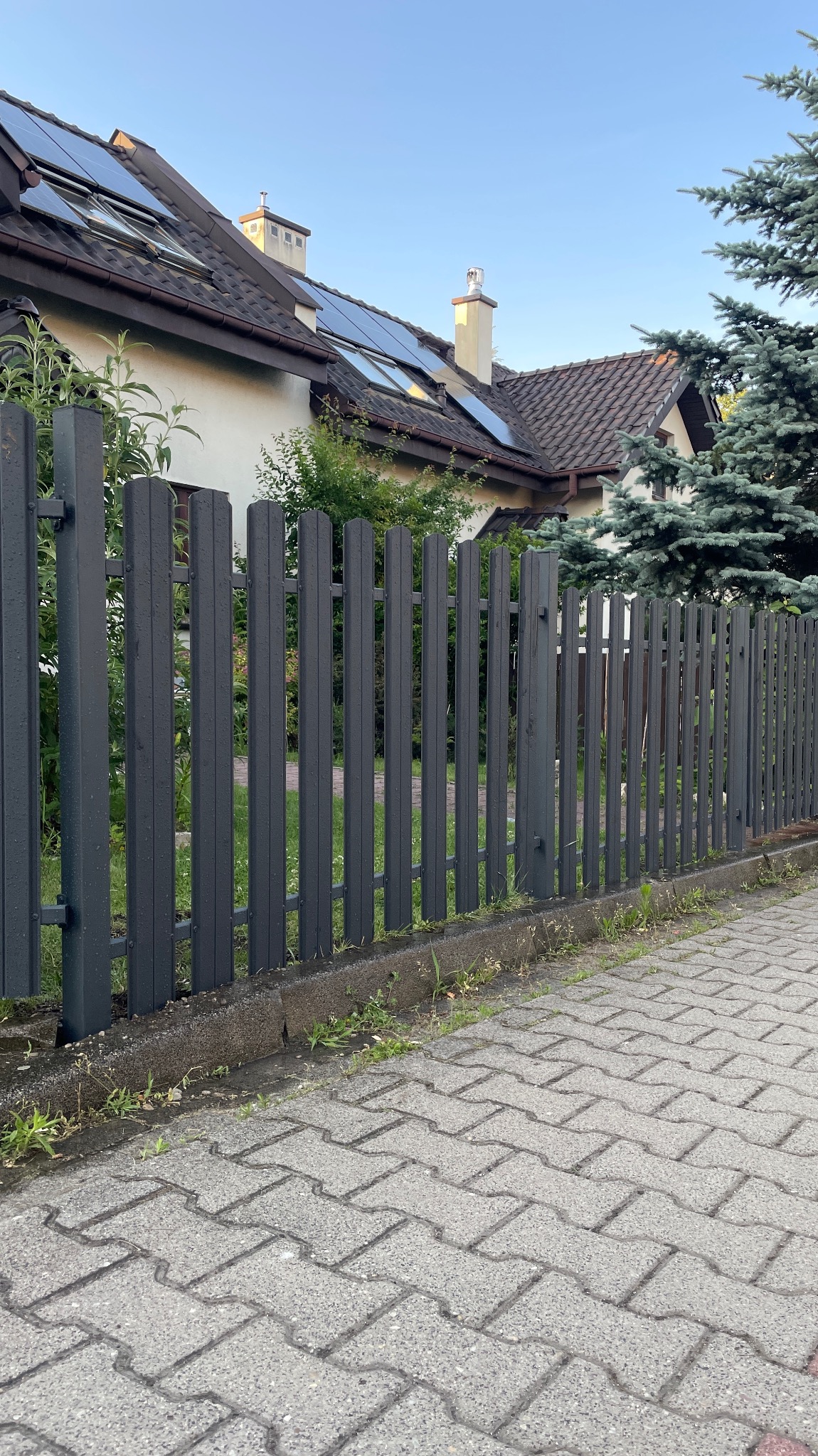 Nowoczesne, grafitowe ogrodzenie palisadowe posesji z domem z panelami słonecznymi w tle, widok z poziomu chodnika z kostki brukowej.