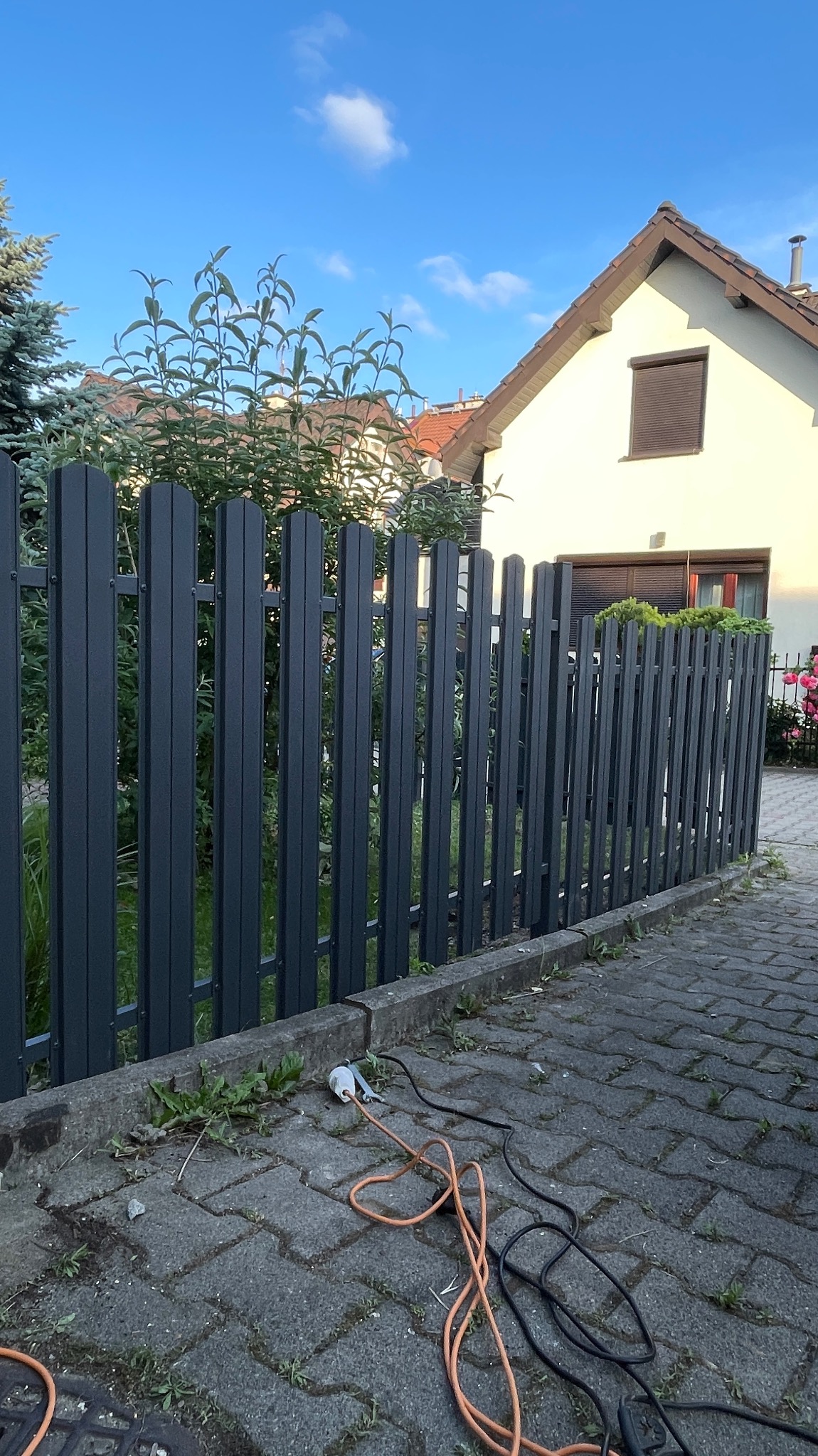 Grafitowe, metalowe ogrodzenie sztachetowe przed domem, na kostce brukowej leżą przewody. Tło: dom z brązową roletą i zielona roślinność.