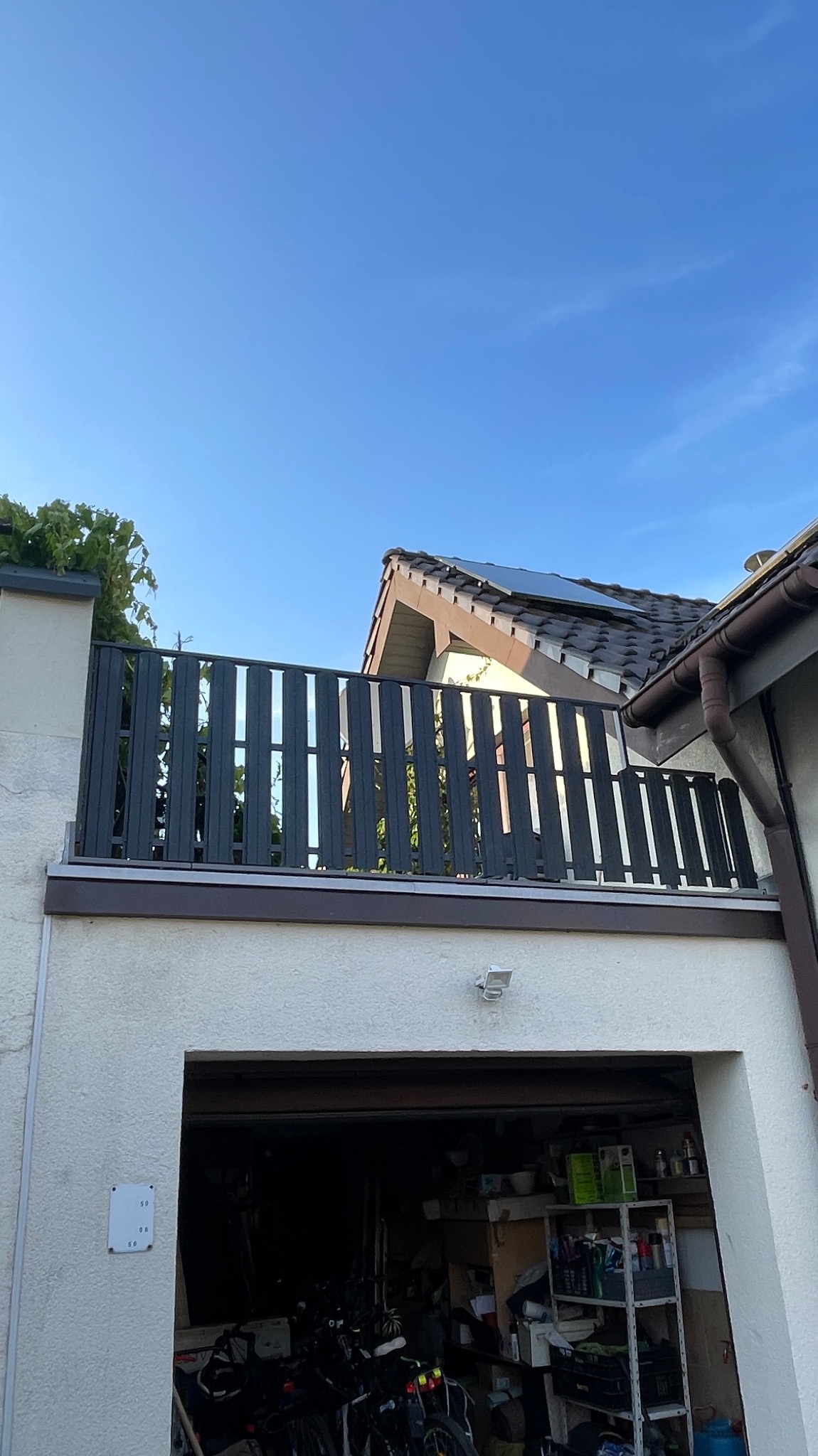 Nowoczesna balustrada balkonowa z pionowych paneli w Trzebini. Szara konstrukcja na białym budynku, widok z dołu na tle błękitnego nieba i fragmentu dachu.