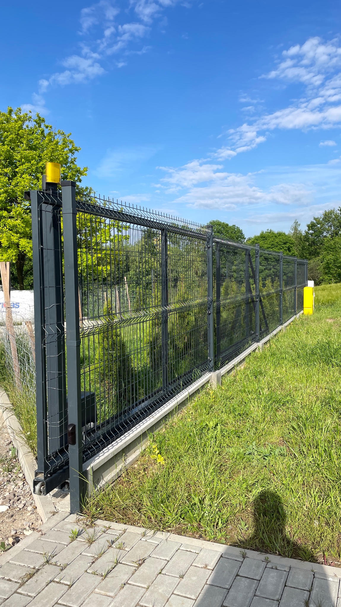 Przesuwna brama panelowa w kolorze grafitowym, z żółtą lampą ostrzegawczą, zamontowana na betonowej podstawie. Widoczne zielone trawniki i drzewa w tle.