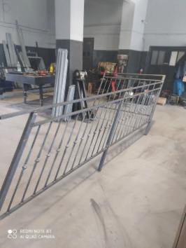 Szara, metalowa balustrada w warsztacie. Widoczne spawy i surowy metal. W tle narzędzia i materiały, co sugeruje etap produkcji lub montażu.